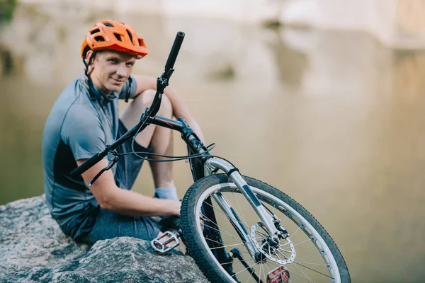 Smiling young trial biker relaxing on rocky cliff over water and looking at camera — Stock Image Smiling Young Trial Biker Relaxing Rocky Cliff Water Looking Camera Stock Image