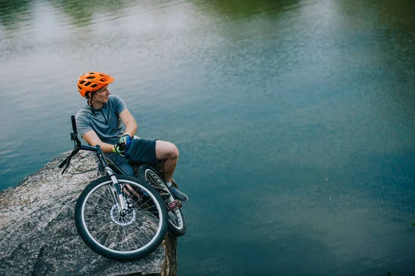 Handsome young trial biker relaxing on rocky cliff over water — Stock Image Handsome Young Trial Biker Relaxing Rocky Cliff Water Stock Image