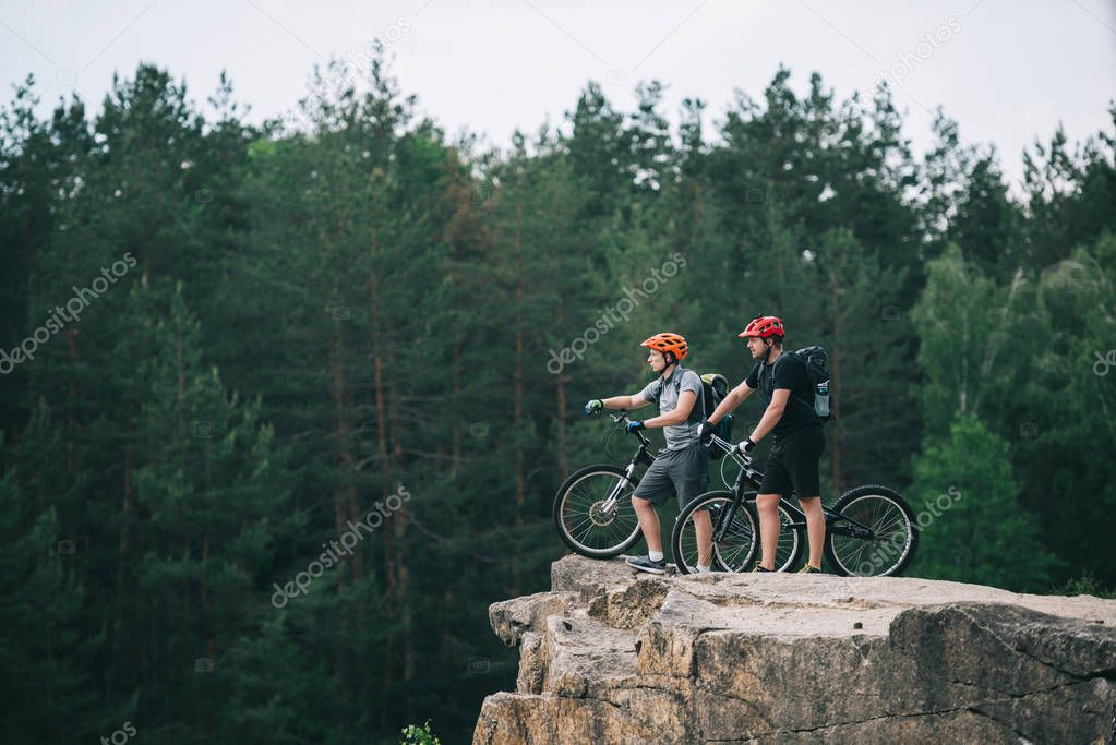 side view of young trial bikers standing on rocky cliff with blurred pine forest on background and pointing somewhere