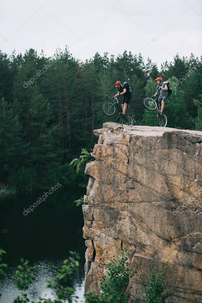 side view of young trial bikers standing on back wheels on rocky cliff over lake with blurred pine forest on background