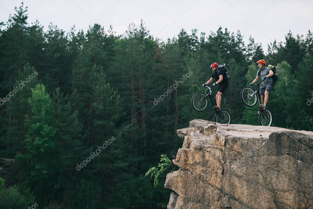 side view of young trial bikers with backpacks standing on back wheels on rocky cliff with blurred pine forest on background