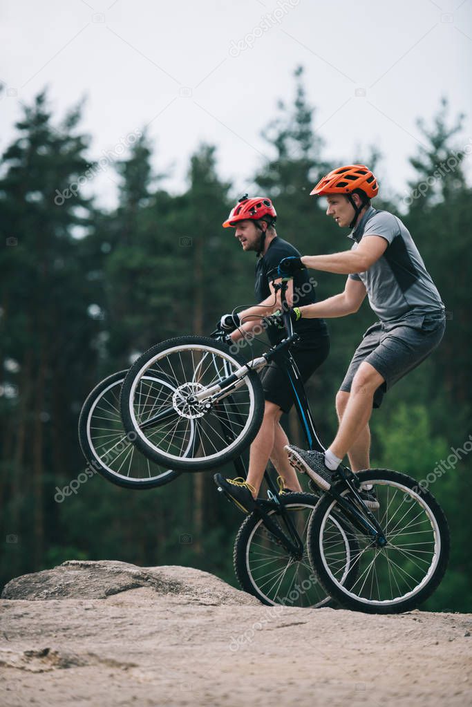 handsome young trial bikers standing on back wheels on rocky cliff with blurred pine forest on background