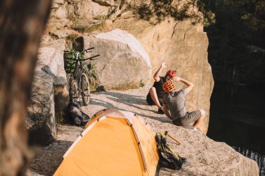high angle view of active trial bikers sitting on rocky cliff with camping tent and giving high five clipart