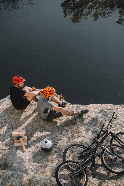 high angle view of young trial bikers eating canned food on rocky cliff