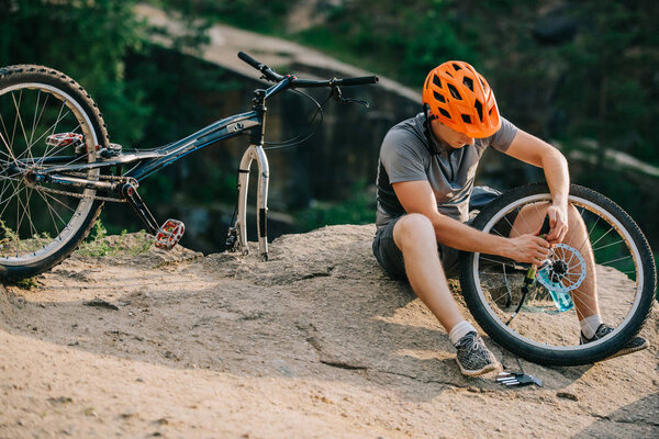 young trial biker fixing bicycle wheel outdoors while sitting on stone