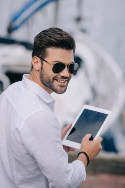 handsome young man in sunglasses smiling at camera while using digital tablet with blank screen clipart