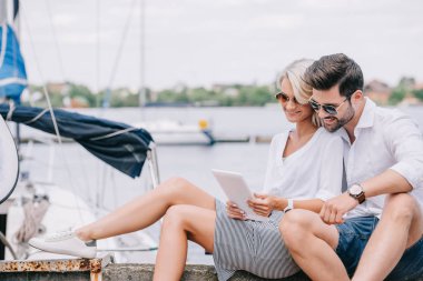 smiling young couple in sunglasses sitting and using digital tablet near yacht clipart