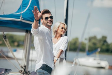 happy young man in sunglasses smiling at camera and waving hand while holding hands with beautiful girl on yacht  clipart