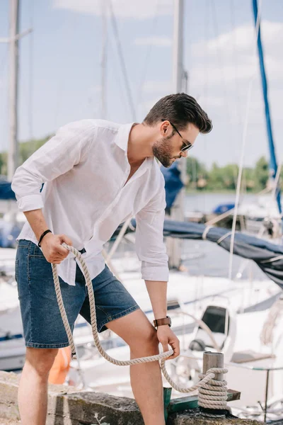 ヨットの近くに立っている間ロープを保持しているサングラスでハンサムな若い男 — ストック写真