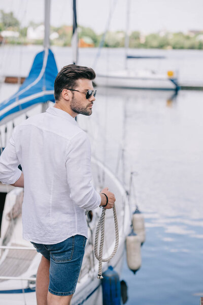 handsome young man in sunglasses looking away while holding rope at yacht