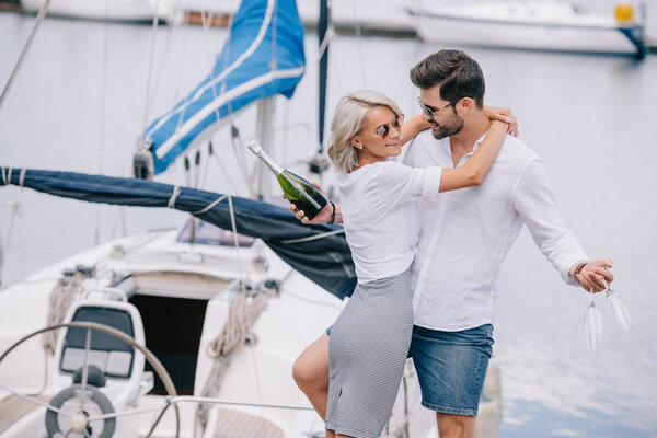 Улыбающаяся девушка в солнечных очках обнимает красивого мужчину с бутылкой вина и бокалами на яхте
