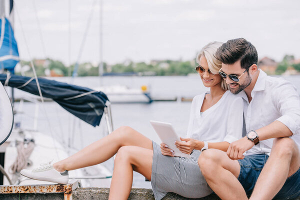 smiling young couple in sunglasses sitting and using digital tablet near yacht
