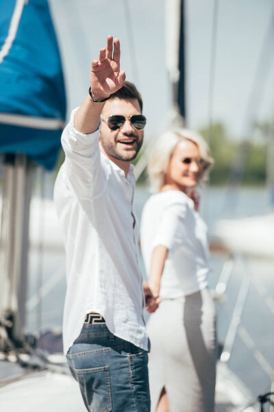 young man in sunglasses smiling at camera and waving hand while holding hands with beautiful girl on yacht 