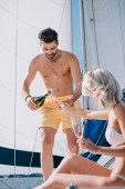 pohledný muž košili v plavky vylévání šampaňského do sklenice přítelkyně na jachtě 