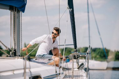 handsome young man in sunglasses looking away while sitting on yacht clipart