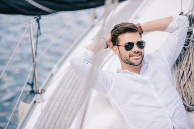 handsome smiling young man in sunglasses resting with hands behind head on yacht clipart