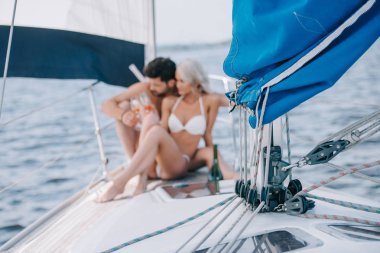 selective focus of sail and couple in swimwear relaxing with champagne glasses behind on yacht  clipart
