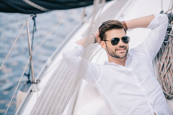Красивый улыбающийся молодой человек в солнечных очках отдыхает с руками за головой на яхте
