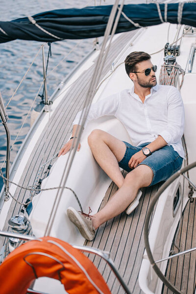 handsome young man in sunglasses looking away while sitting on yacht
