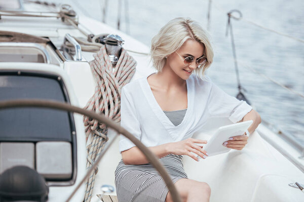 smiling blonde girl in sunglasses using digital tablet while sitting on yacht