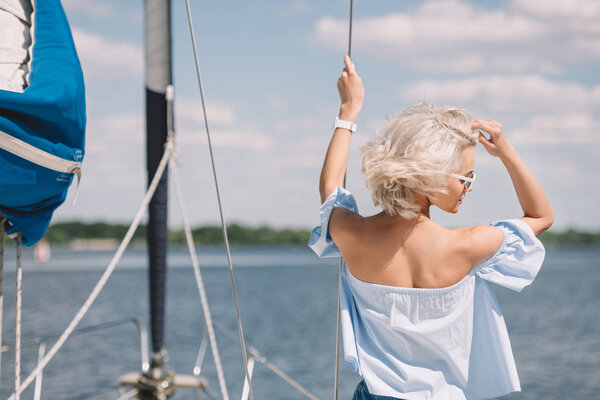 rear view of beautiful young blonde woman in sunglasses looking away while standing on yacht 