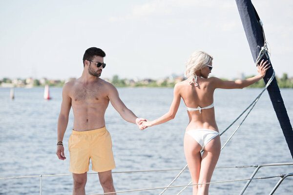 young couple in swimwear and sunglasses holding hands while standing together on yacht