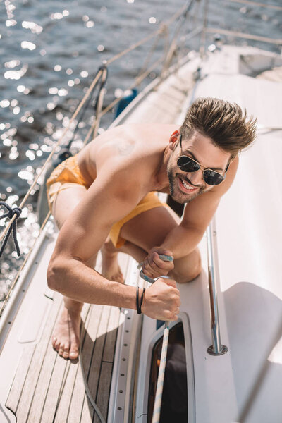 smiling shirtless man in sunglasses pulling rope on yacht 