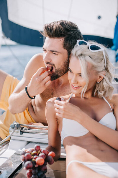 smiling young couple in swimwear eating grapes on yacht 