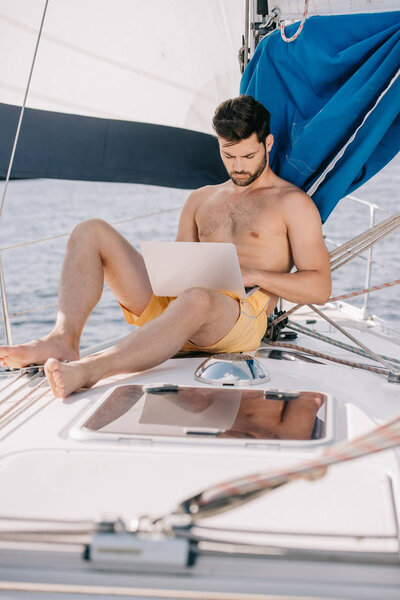 focused shirtless man in swim trunks using laptop on yacht