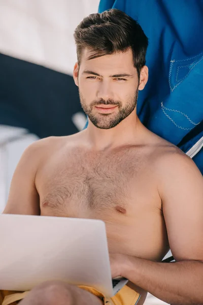 smiling shirtless man in swim trunks using laptop on yacht