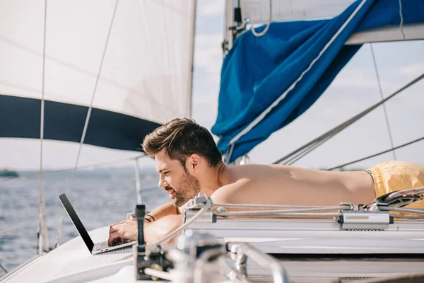 在游艇上使用笔记本电脑的微笑裸男男子侧面视图 — 图库照片