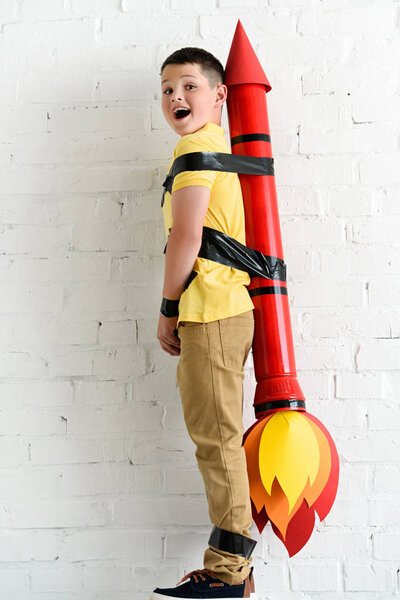 smiling pre-adolescent boy standing with rocket toy on back at home