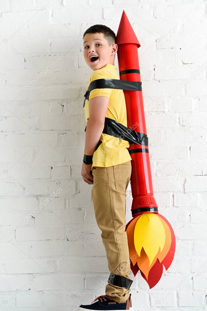 smiling pre-adolescent boy standing with rocket toy on back at home
