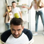 Tied father sitting on floor with apple on head for target at home, parenthood concept