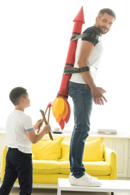 disobedient son playing with tied father with rocket toy on back at home clipart