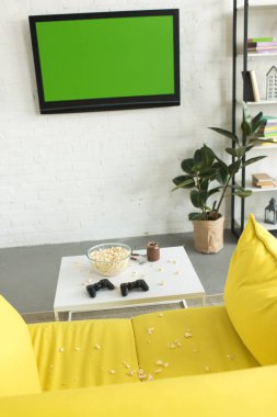 high angle view of glass bowl with popcorn and joysticks on table near yellow sofa in living room clipart