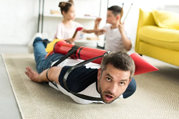 tied father with rocket toy lying on floor and children playing in living room