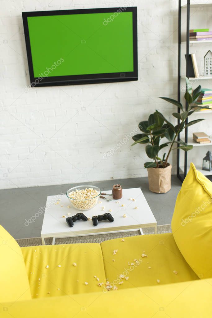 high angle view of glass bowl with popcorn and joysticks on table near yellow sofa in living room