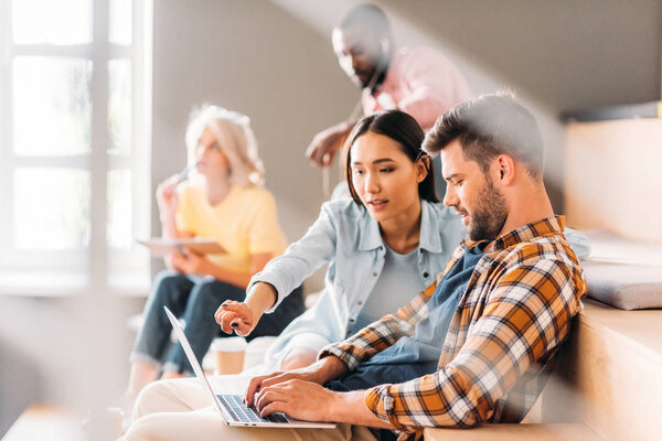 боковой вид молодых студентов, использующих ноутбук, сидя на трибунах вместе в колледже
