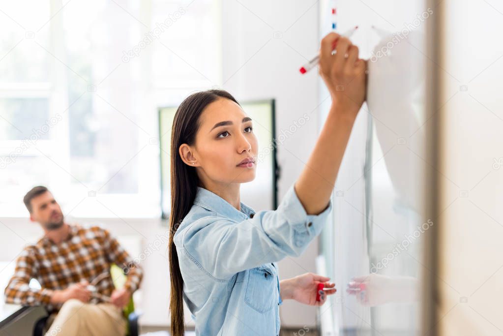 young asian student girl writing on whiteboard during lesson with blurred teacher on background