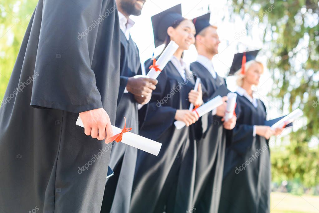 cropped shot of graduated students in capes and hats holding rolled diplomas