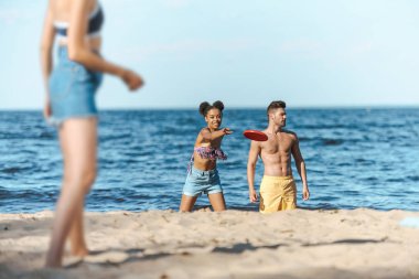 selective focus of interracial friends playing with flying disk on sandy beach clipart