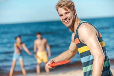 selective focus of smiling young man holding flying disk with friends behind on sandy beach clipart
