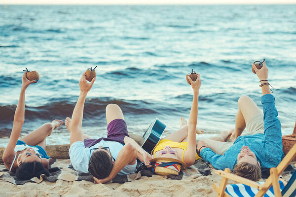 multicultural friends with coconut cocktails and acoustic guitar resting on sandy beach