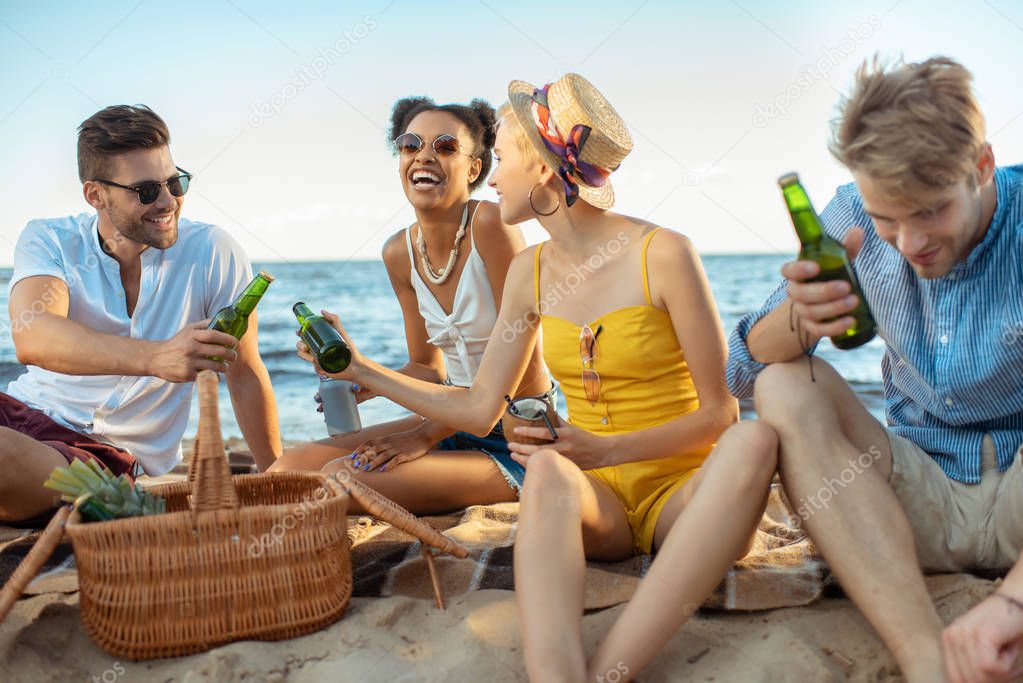 interracial smiling young friends with beer resting on sandy beach together on summer day