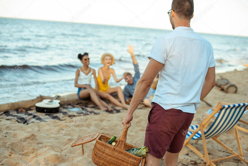 selective focus of man with picnic basket and multiethnic friends on sandy beach