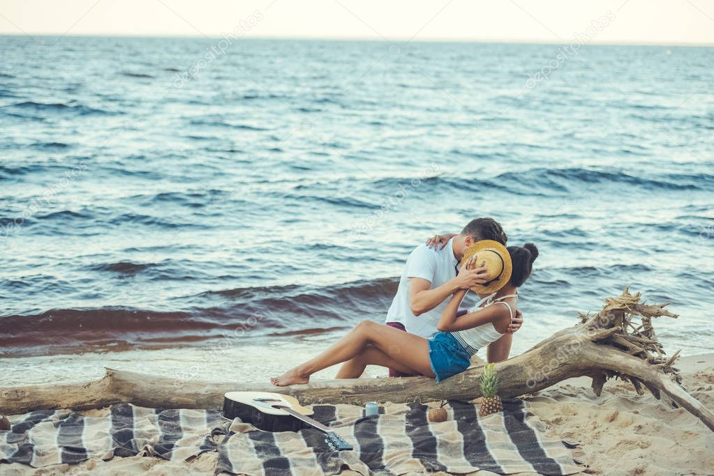 obscured view of multiethnic couple hiding behind hat while kissing on beach