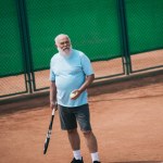 Anciano en ropa deportiva con raqueta de tenis y pelota de pie en la cancha