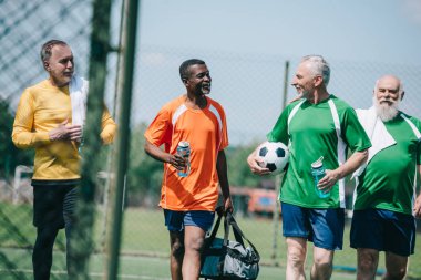 group of interracial elderly sportsmen with sportive water bottles walking on football field clipart
