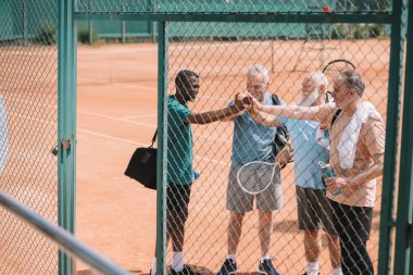 multicultural group of elderly tennis players holding hands together after game on court clipart